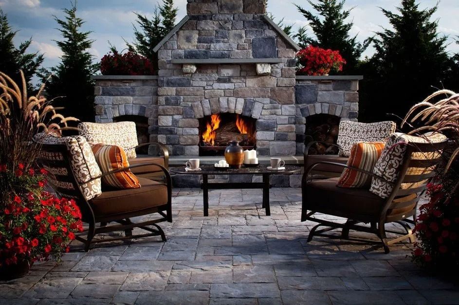 Stone outdoor fireplace with two cushioned patio chairs and flower pots in a cozy backyard setting
