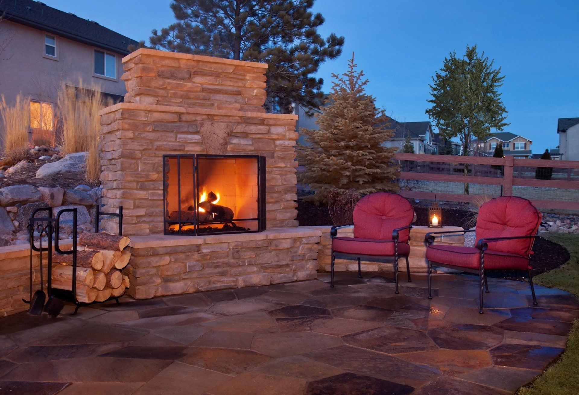 Stone outdoor fireplace on a patio at dusk, with stacked firewood and two red chairs nearby.