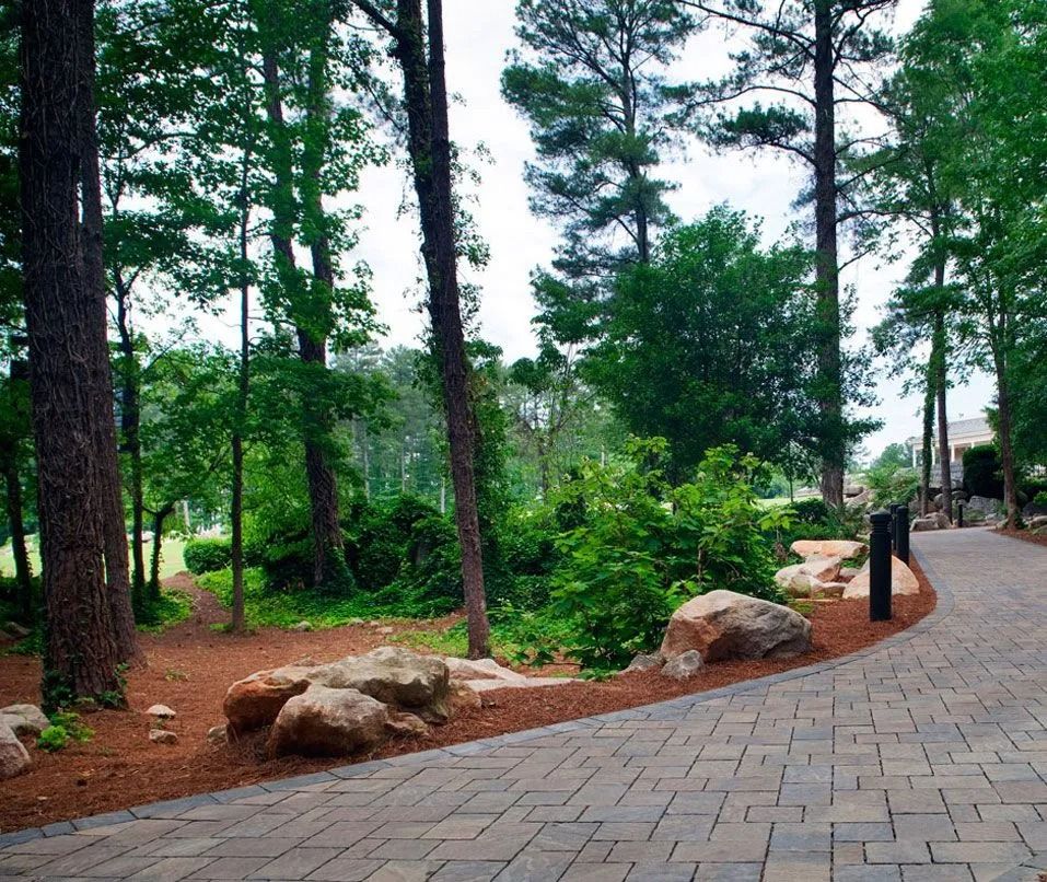 Tree-lined park path with brick paving, rocks, and green shrubs beside a quiet roadway.