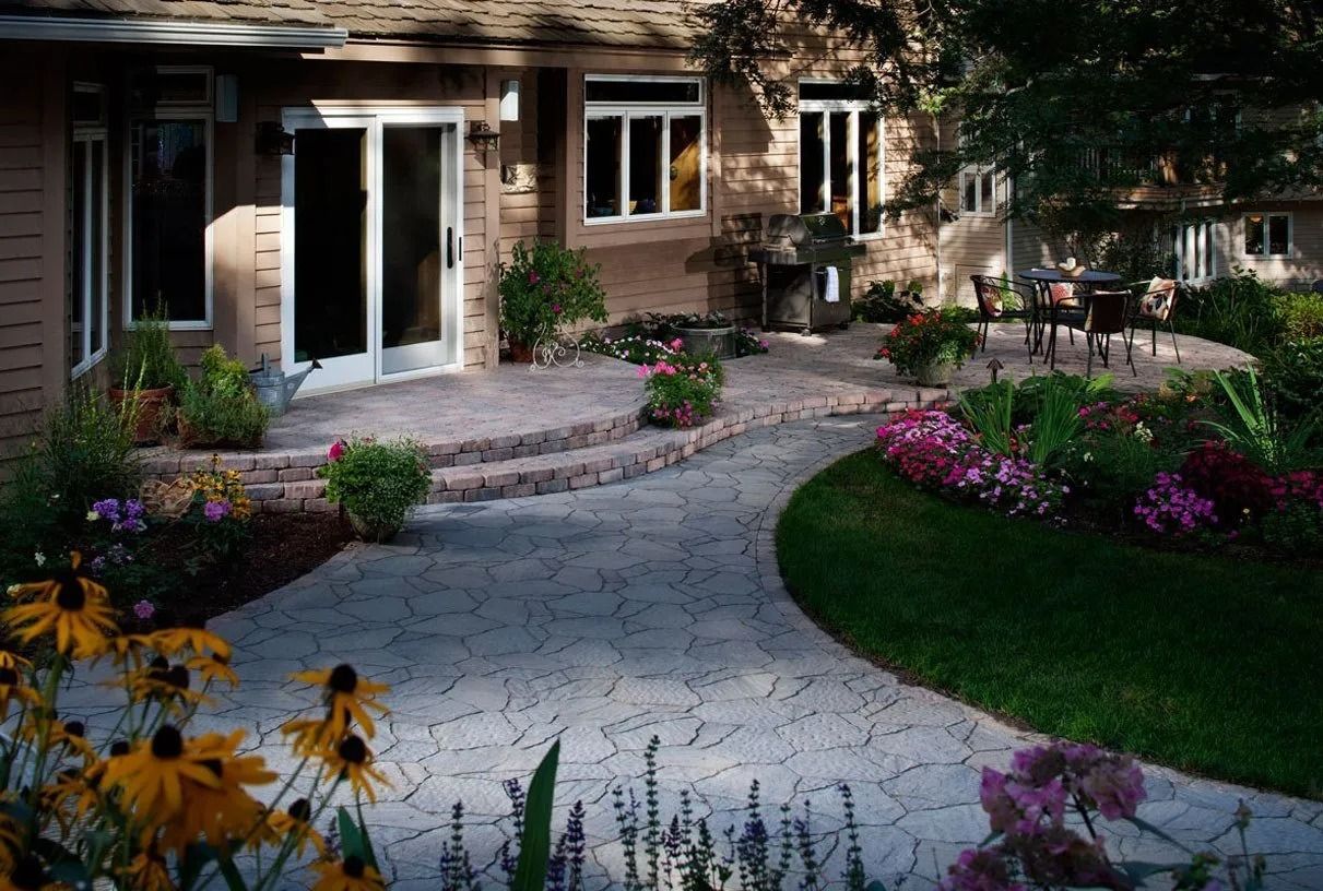Brick house patio with curved stone path, flower beds, and outdoor seating in a landscaped garden.