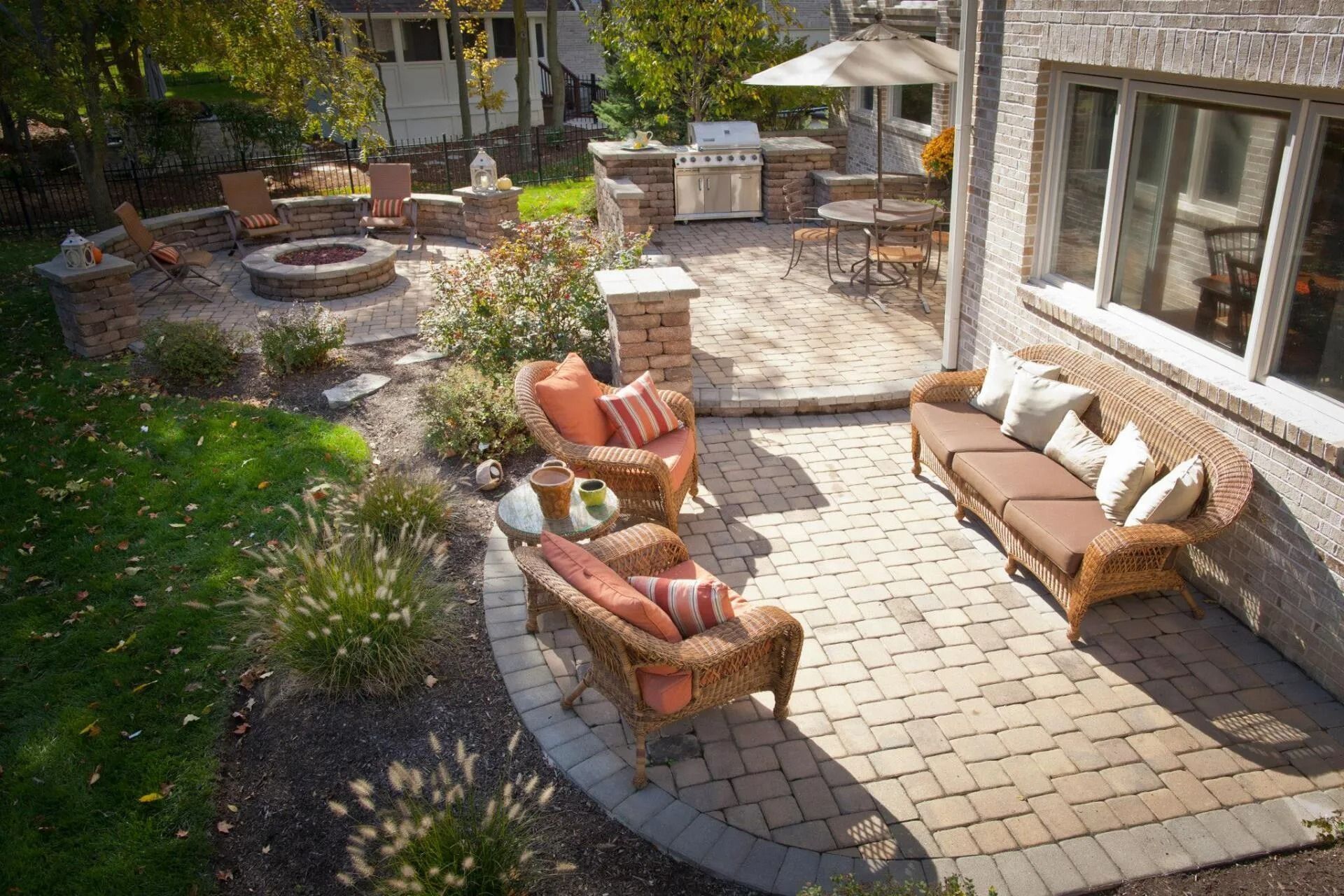 Sunlit backyard patio with wicker seating, orange cushions, potted plants, and a stone fire pit