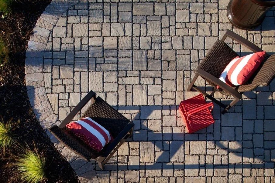 Two wicker chairs with red-and-white cushions on a stone patio, separated by a red folding stool