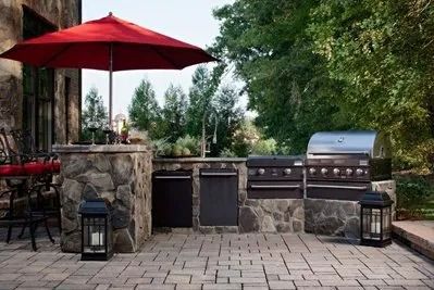 Outdoor patio with stone kitchen, grill, red umbrella, and lanterns on a paved terrace