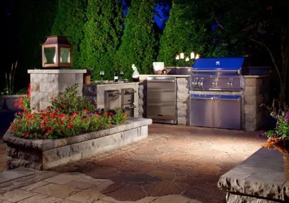 Stone patio with a flower bed, lanterns, and a blue stainless steel grill at night