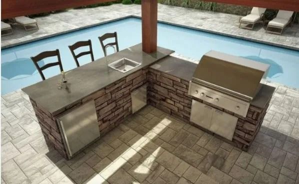 Outdoor kitchen with stone counters, built-in grill, sink, and bar stools beside a pool