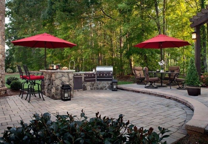 Stone patio with red umbrellas, outdoor seating, and a built-in grill in a wooded backyard.