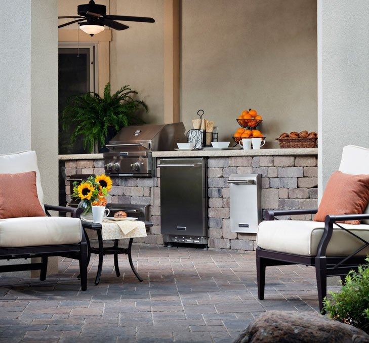 Patio seating area with cushioned chairs, grill, stone bar, and potted flowers in a landscaped backyard