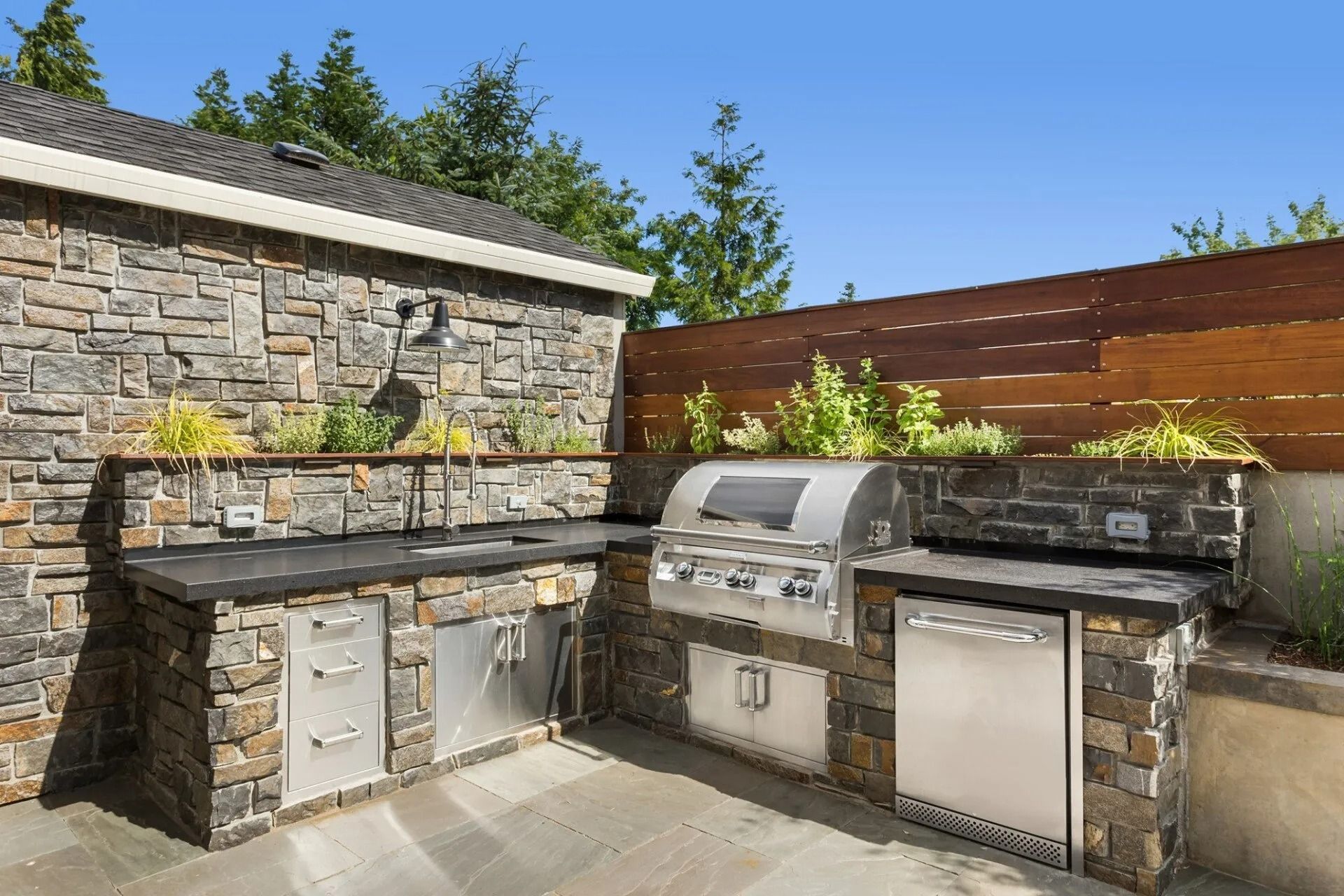 Outdoor stone patio with dual stainless steel grills and cabinets beside a stone wall and wooden fence