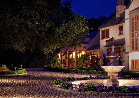 Nighttime courtyard of a lit house with cobblestone path, trees, and a fountain