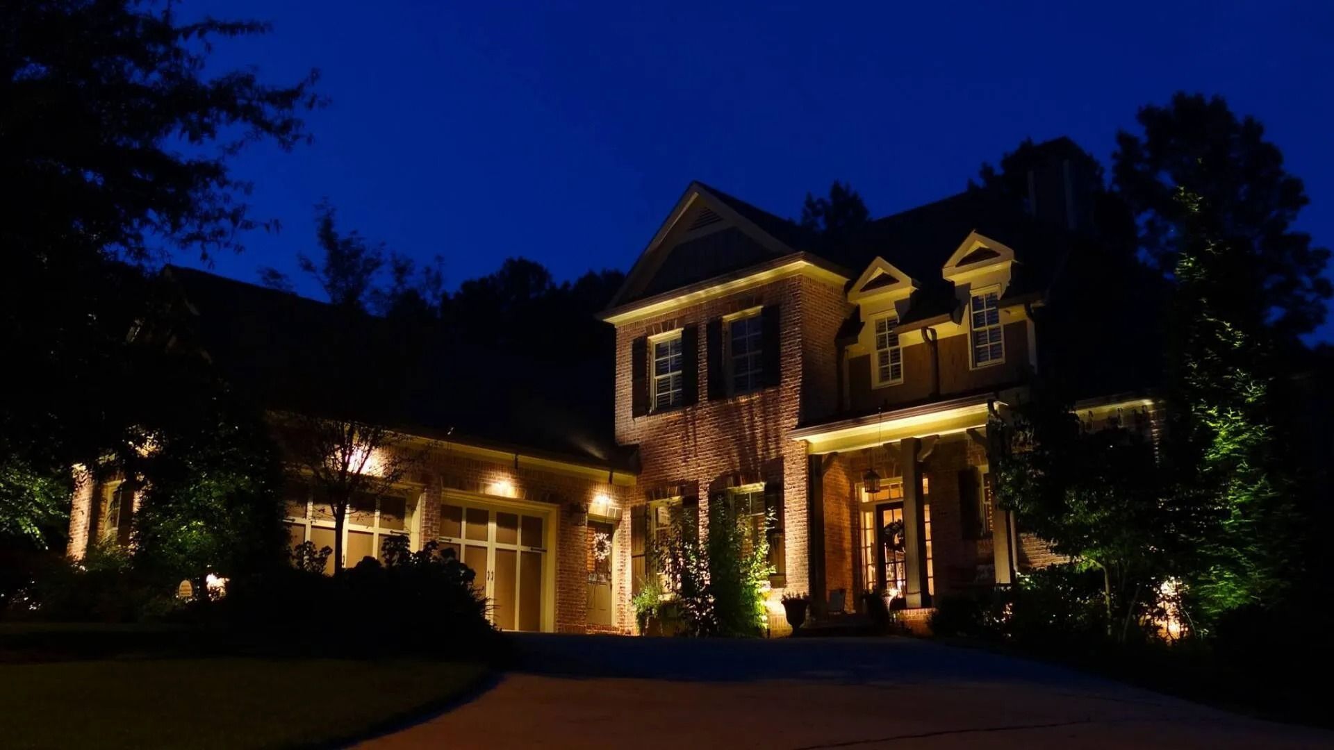 Illuminated brick house at night with warm porch lights and dark trees surrounding it