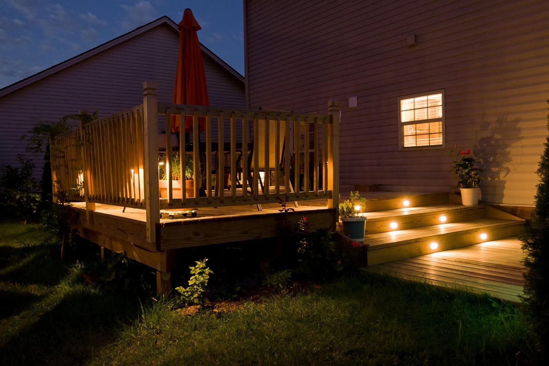 A lit wooden deck and steps beside a house at dusk, with warm outdoor lights and potted plants.