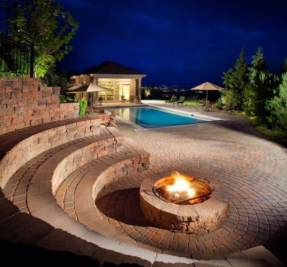 Luxury backyard patio with curved stone seating, fire pit, pool, and lit house at night