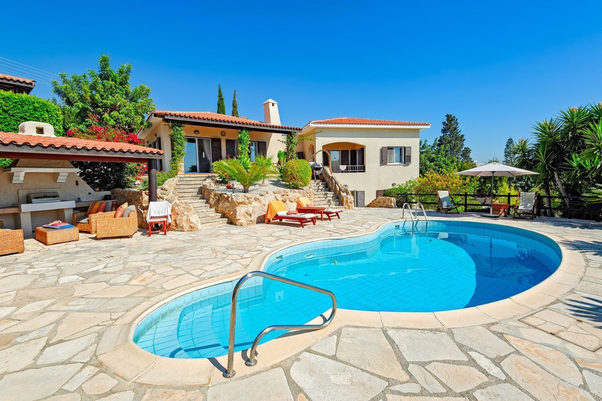 Sunny backyard patio with a blue swimming pool and stone paving beside a house and lounge chairs