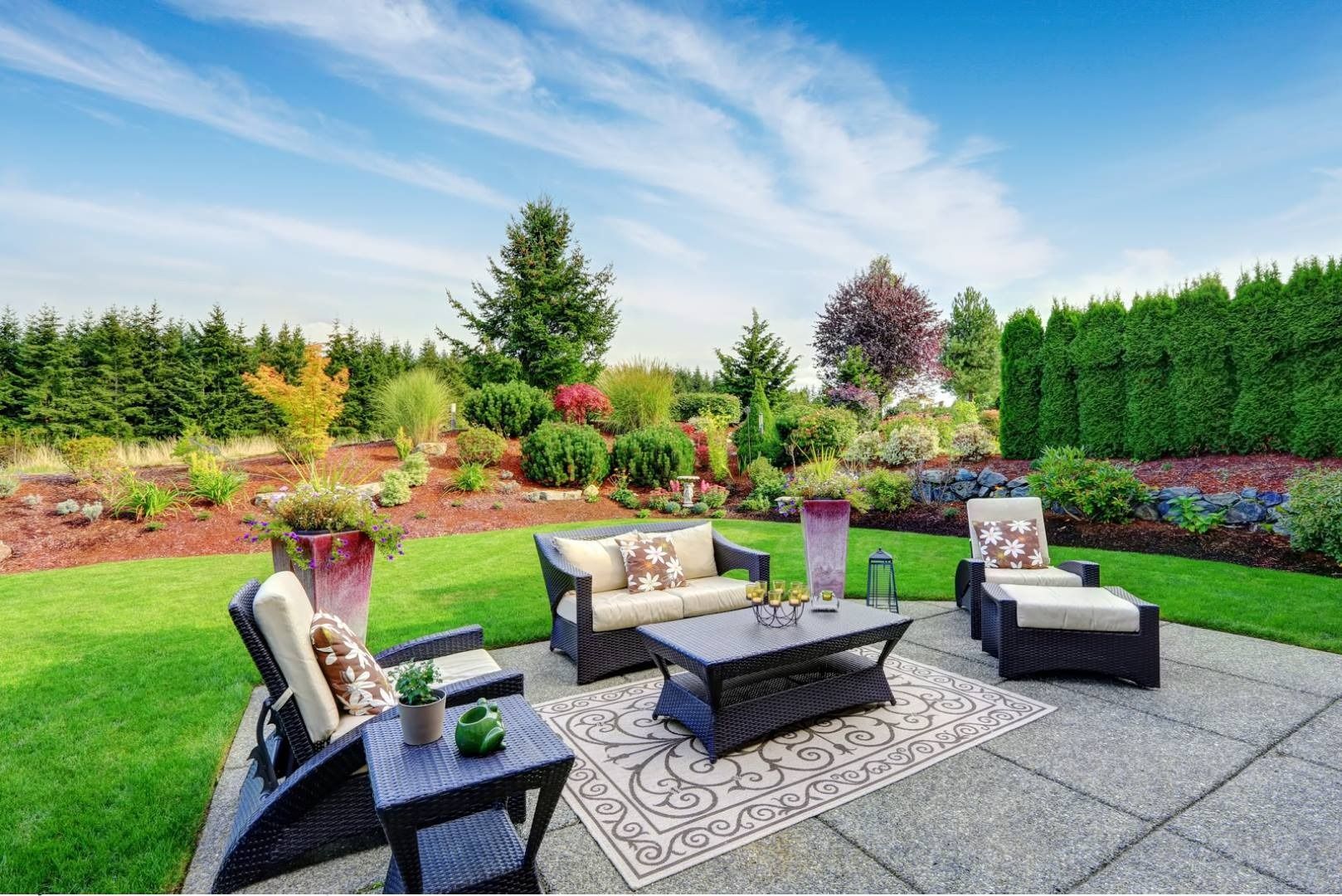 Patio seating area with outdoor chairs and sofa on a patterned rug, surrounded by a lush garden and lawn.