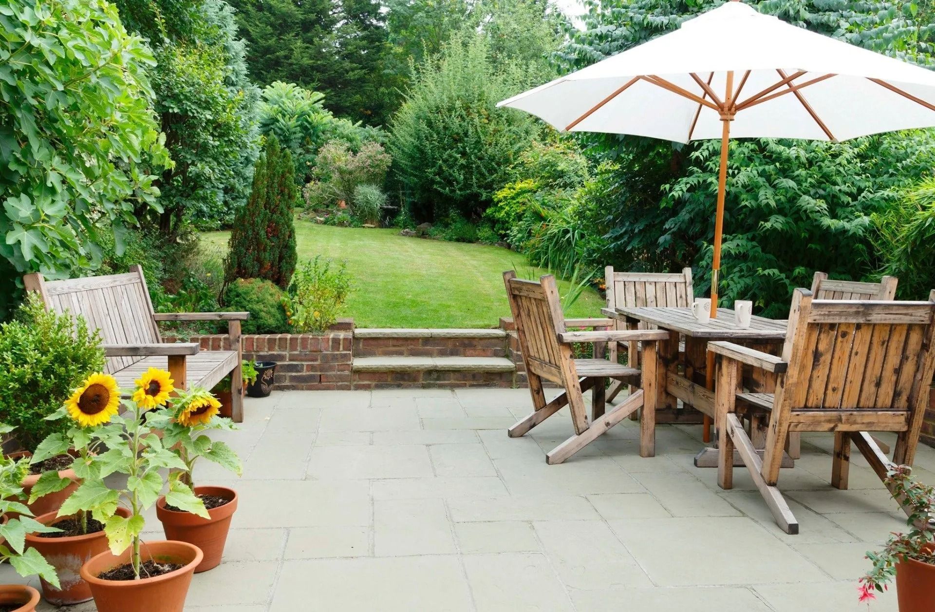 Patio with wooden table and chairs under white umbrella, potted sunflowers, and a lush green garden backdrop