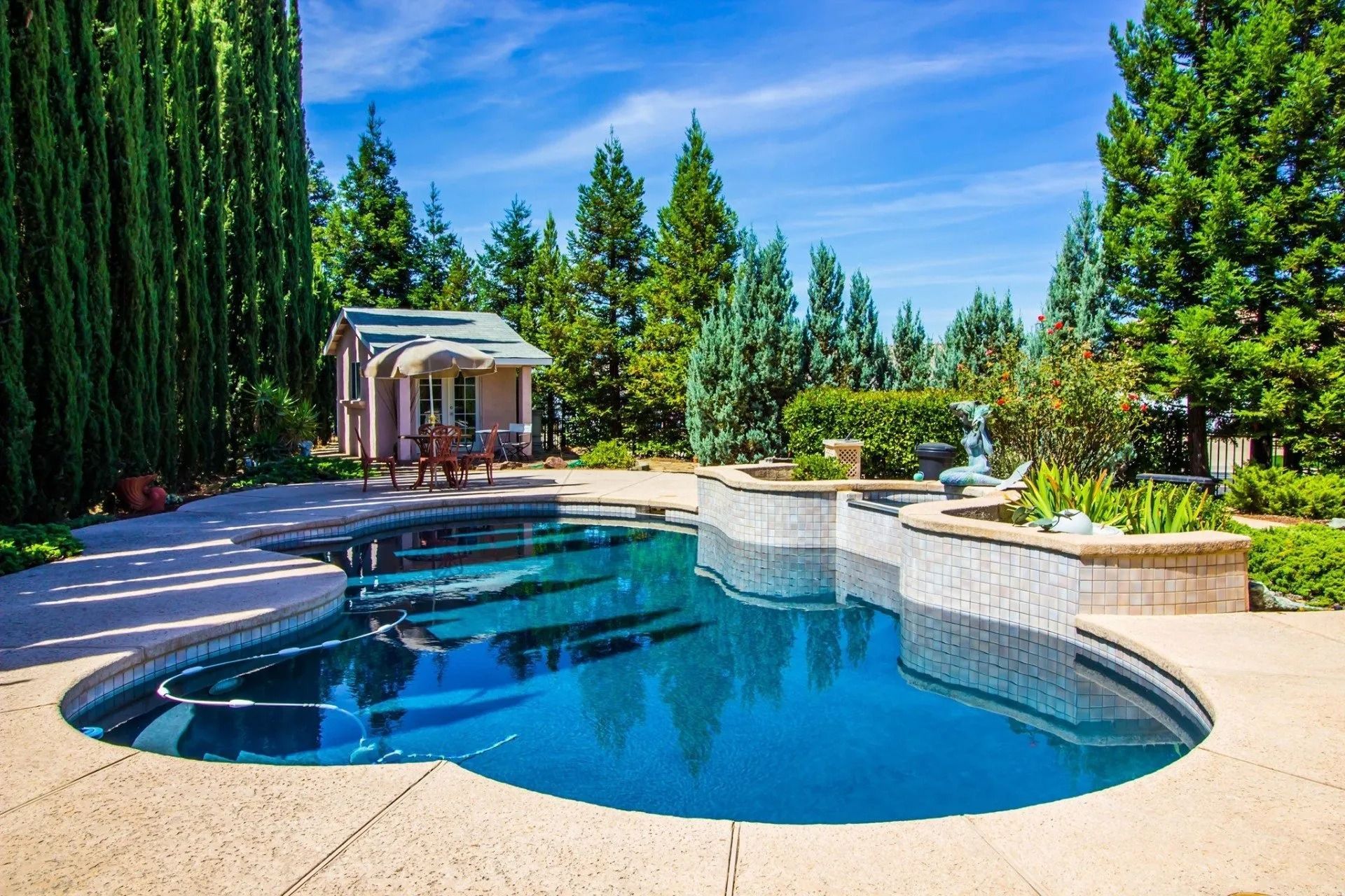 Backyard pool with a stone patio, tall trees, and a small gazebo under a blue sky