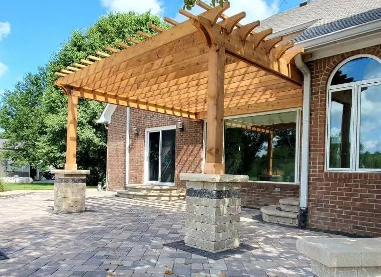 Brick patio with a wooden pergola and stone pillars beside a house with large windows
