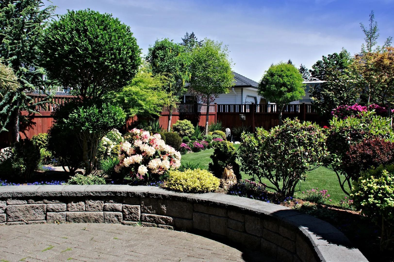 Landscaped garden with flowering shrubs, trees, and a stone retaining wall in front of a house