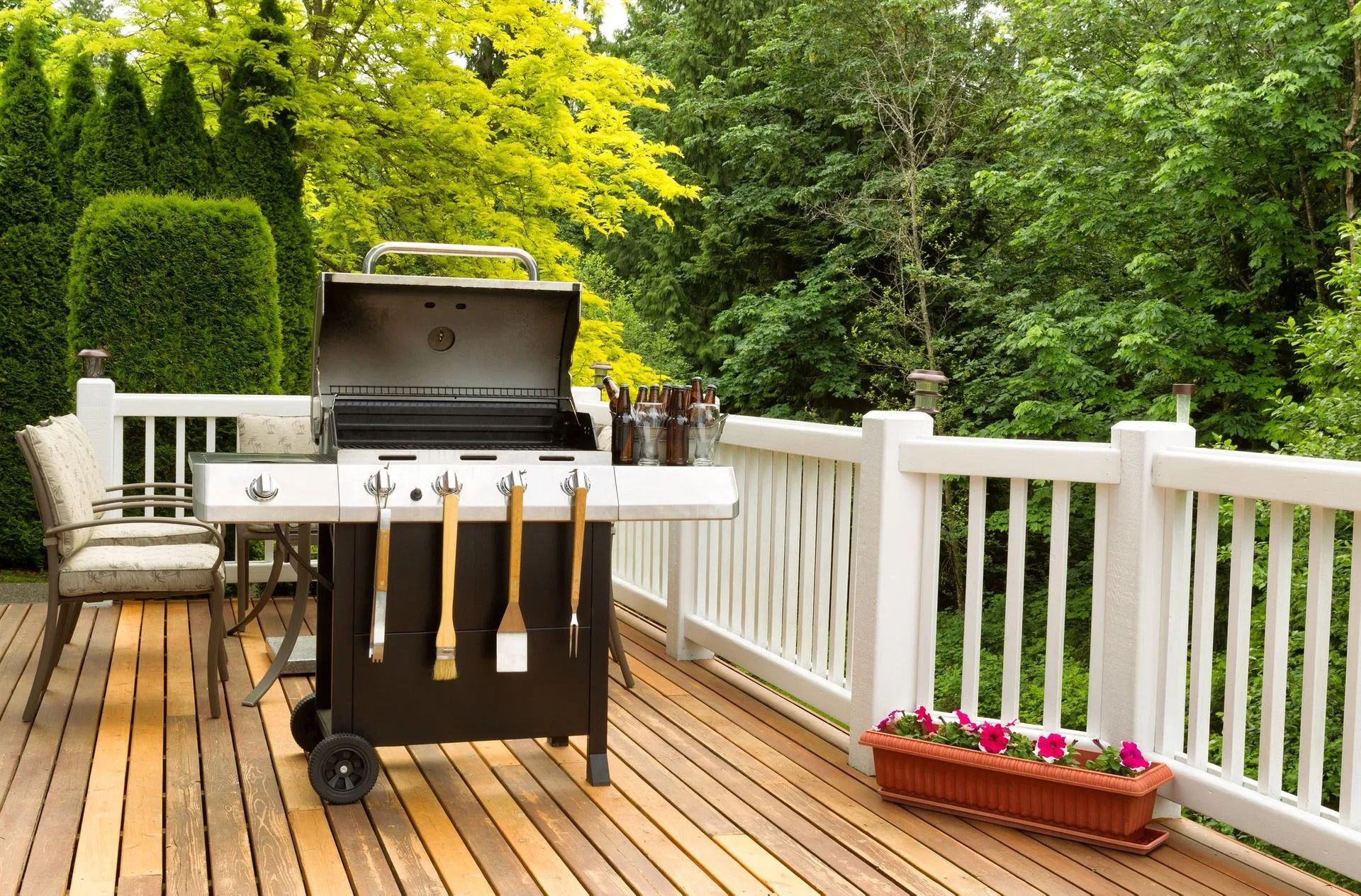 Backyard deck with a black grill, white railing, and flower planter surrounded by trees