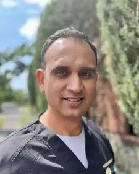 Man in black scrubs smiles outdoors with greenery in the background.