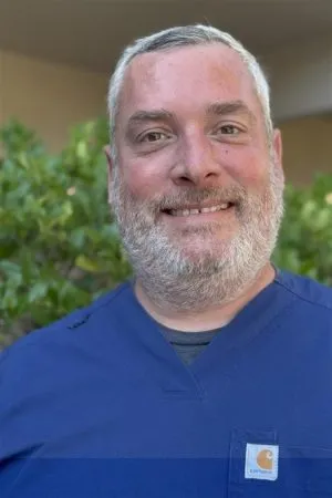 Man with graying beard and blue scrubs smiling, outside near green foliage.