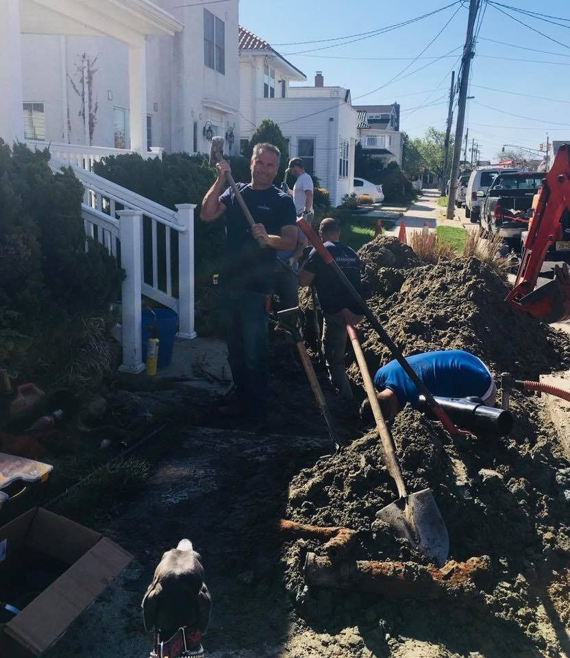 Workers digging a muddy trench beside homes, with shovels and a backhoe on a sunny street.