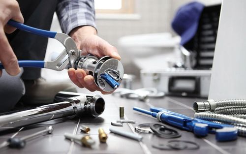 Hands using pliers to work on a metal part on a cluttered workbench with tools and screws
