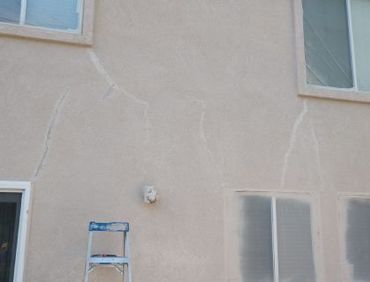Beige stucco exterior wall with several cracks and a blue ladder