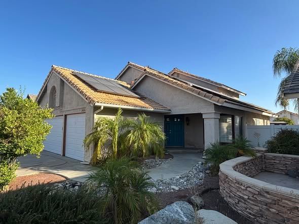 Tan house with solar panels on the roof, two-car garage, and blue front door; sunny day.