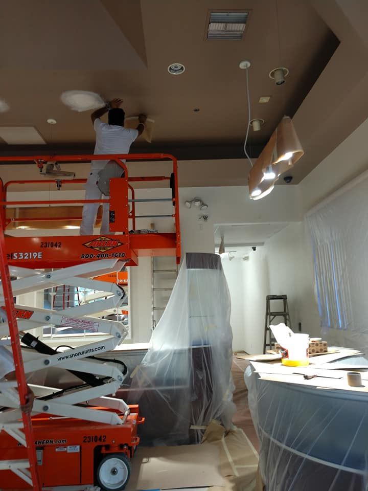 A person on a lift repairs ceiling fixtures in a room covered in plastic, likely during a renovation.
