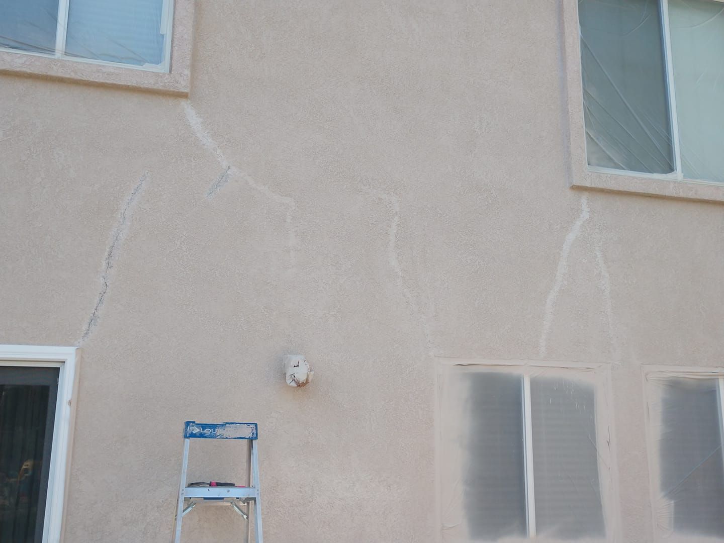 Tan stucco wall with several long, vertical cracks; a small ladder is at the bottom.