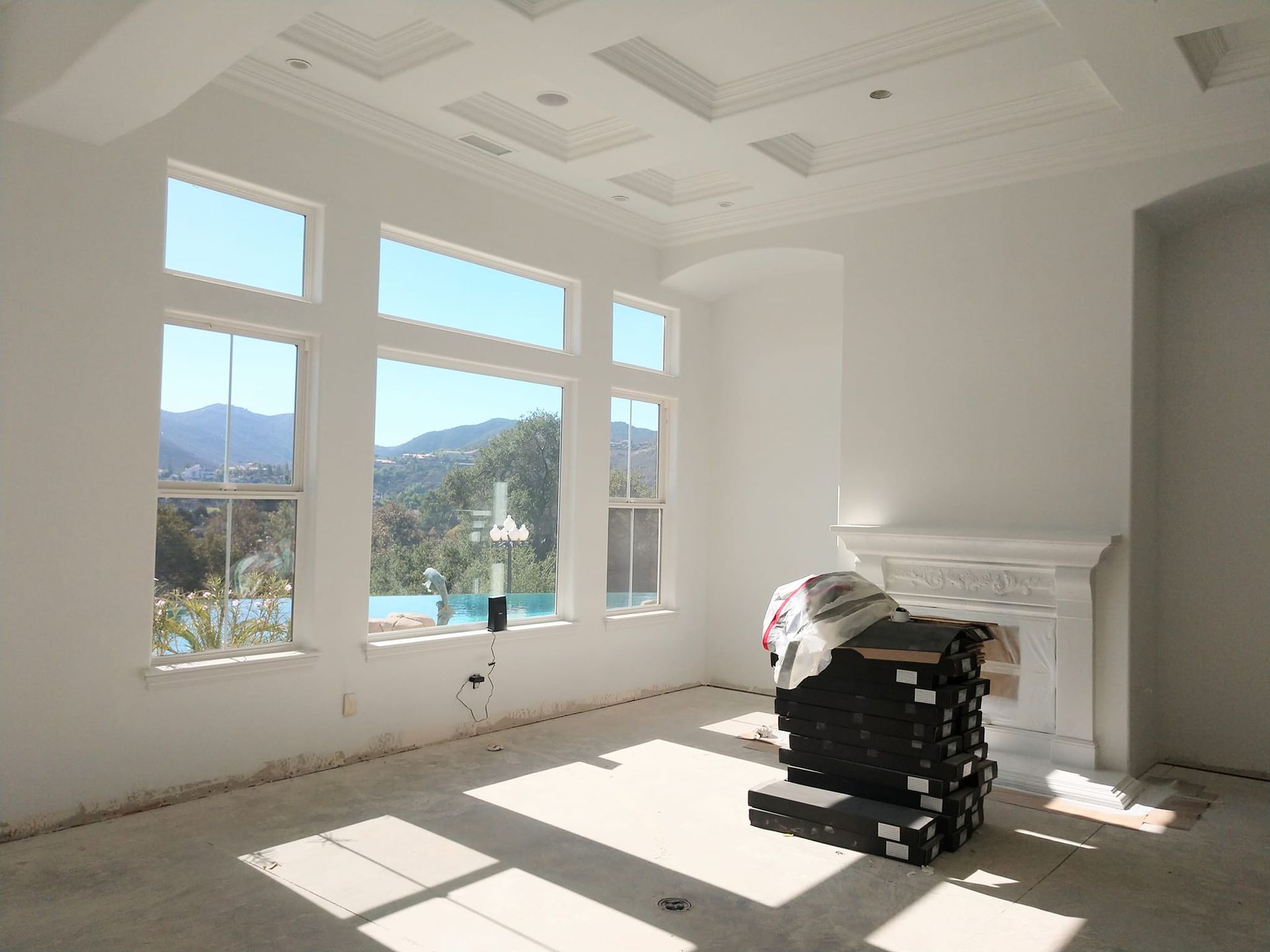 Empty room with large windows overlooking a mountainous landscape. White walls, fireplace, and sunlight streaming in.