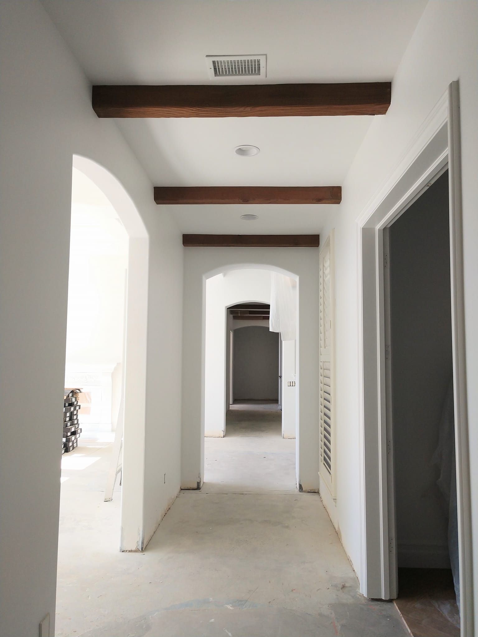 White hallway with arched doorways, dark wooden beams, and concrete floor.