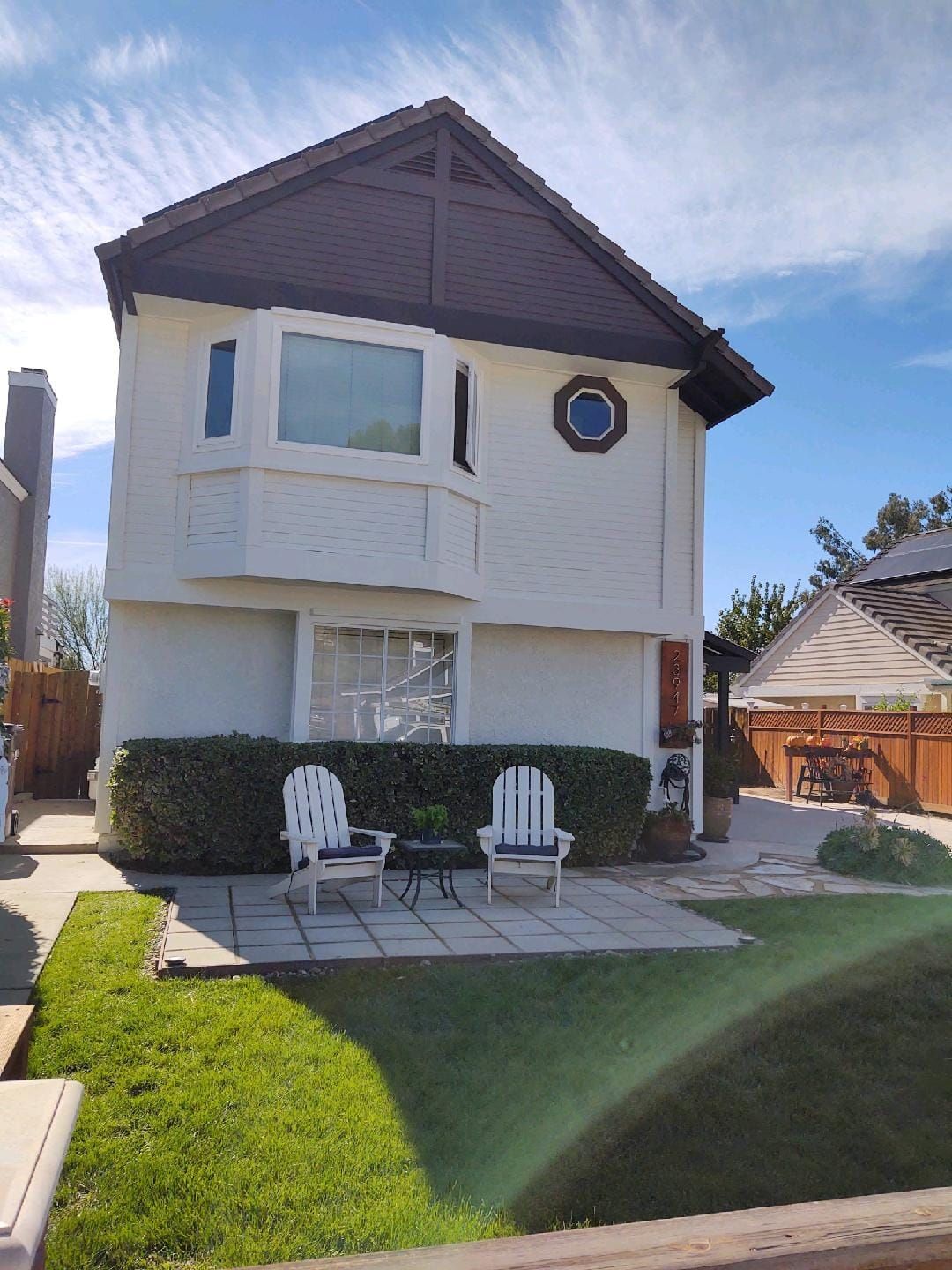 Two-story white house with brown roof and trim, bay window, round window, chairs on patio, green grass, sunny day.