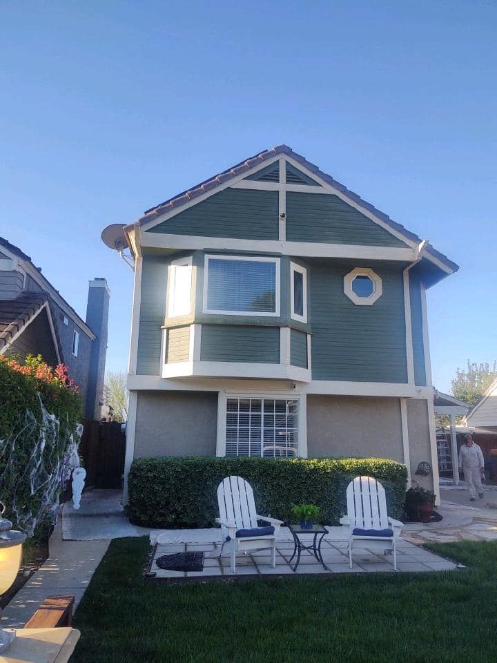 Two-story house with green siding, white trim, two white chairs, and a small table on the lawn.