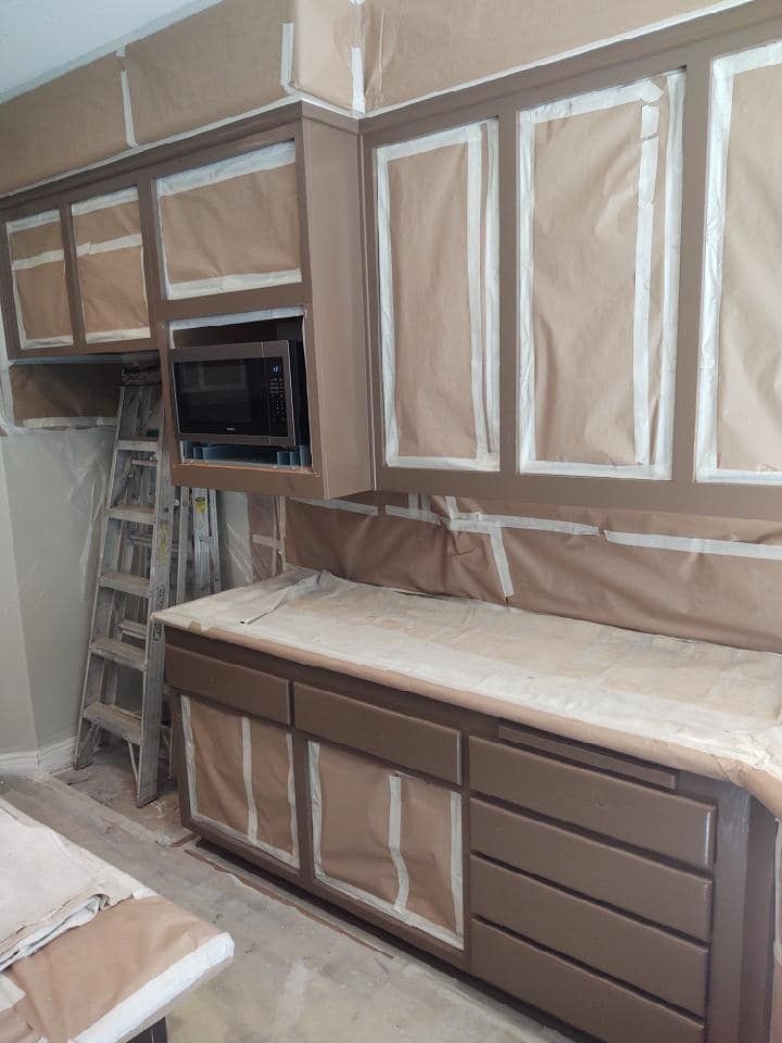 Kitchen cabinets, countertop and microwave oven covered in brown paper and painter's tape, ready for painting.
