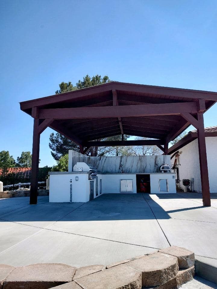 Outdoor kitchen under a brown-roofed pergola. White counter with built-in grill, cabinets. Concrete patio, blue sky.