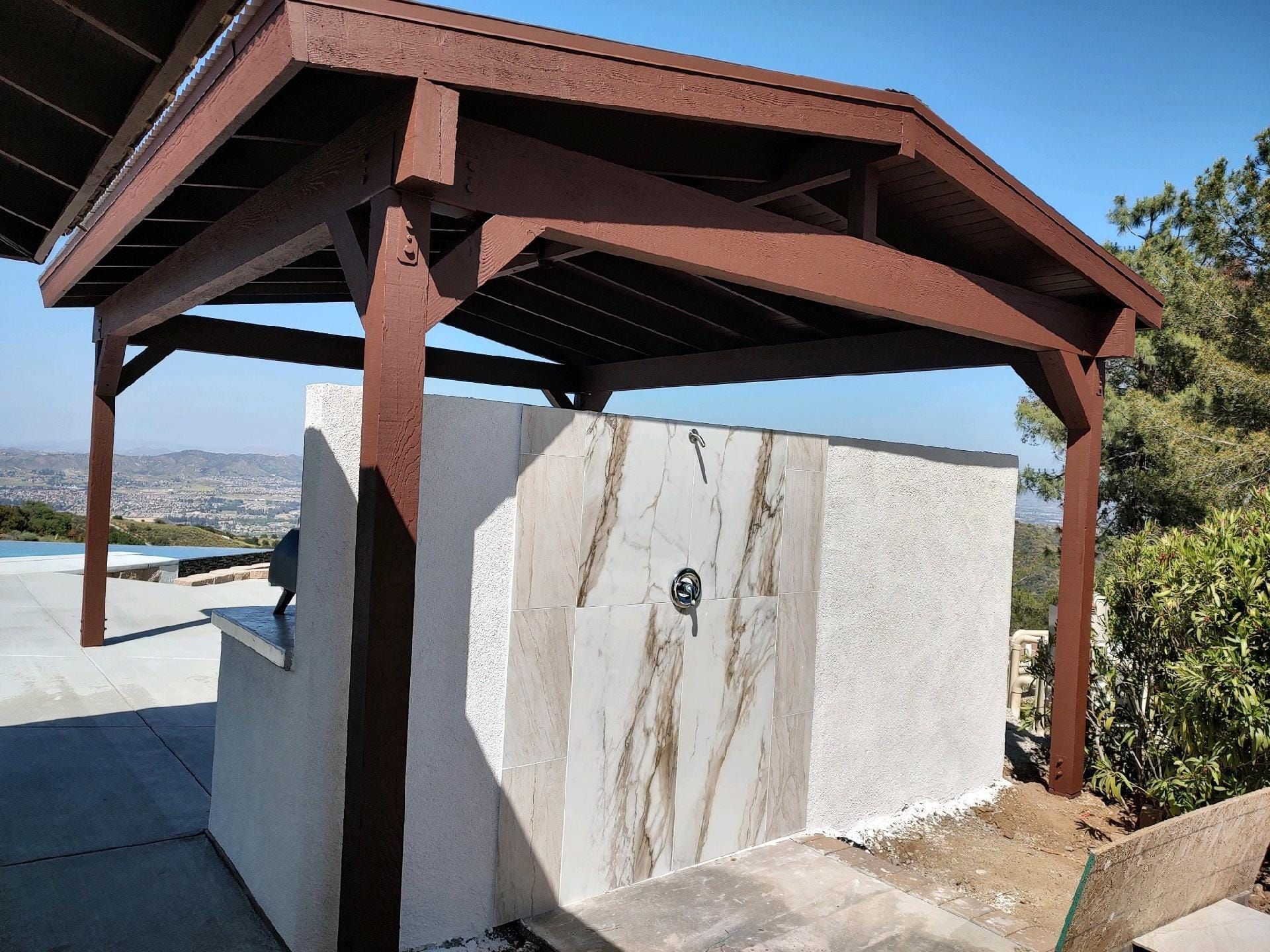 Outdoor shower under brown pergola with city view.