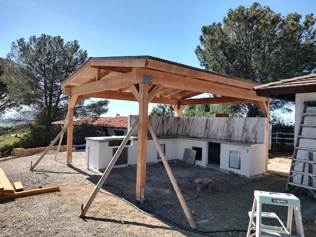 Wooden pergola construction over a partially built outdoor kitchen on a gravel surface.