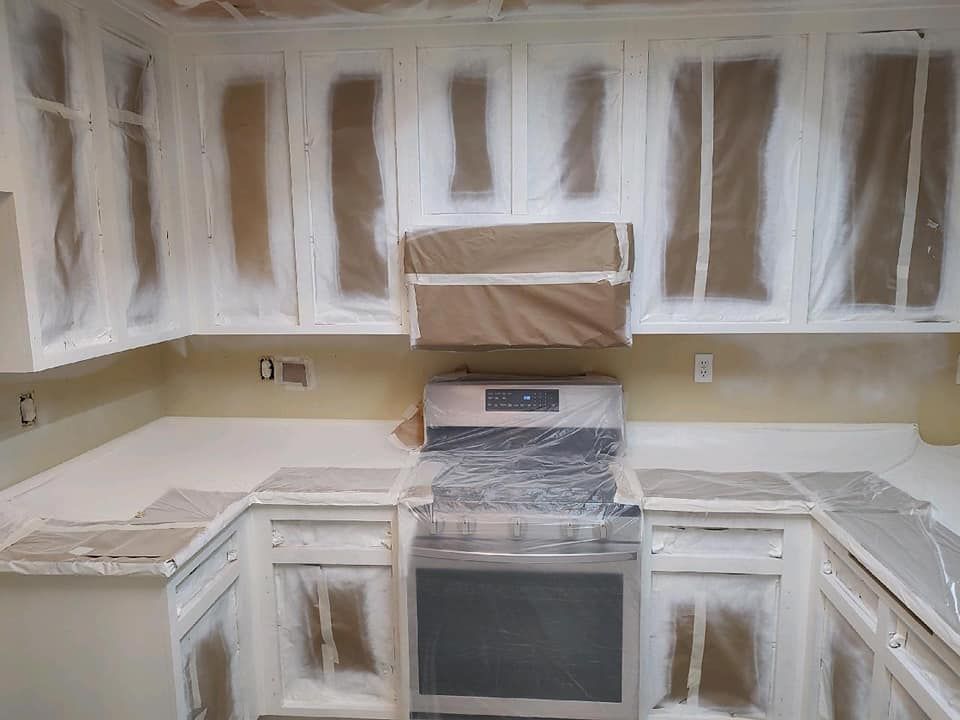 Kitchen cabinets covered in plastic and tape, ready for painting; stove in center.