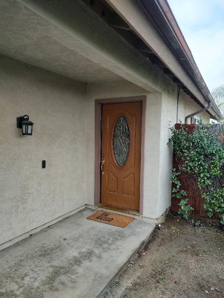 Brown front door with oval glass, under a tan awning. Exterior wall has a black light and a welcome mat.