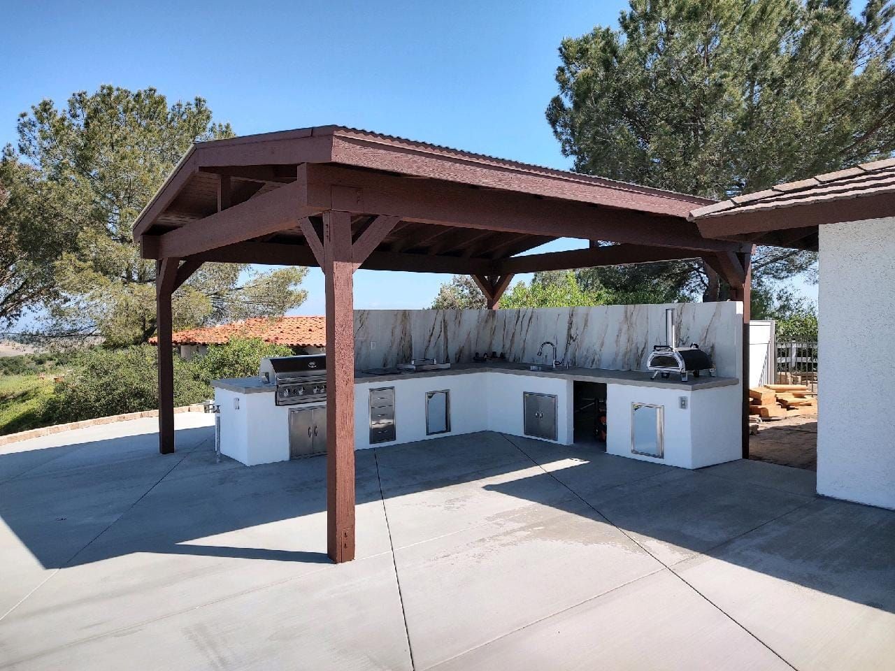 Outdoor kitchen with wooden pergola, white counters, built-in appliances, and a concrete patio.
