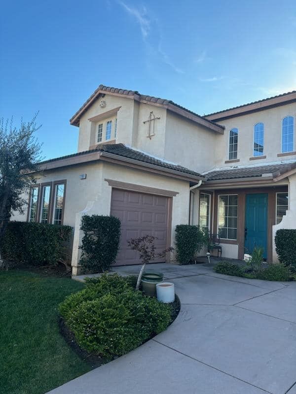 Two-story beige house with a blue door and a brown garage door. Green bushes and grass in front.