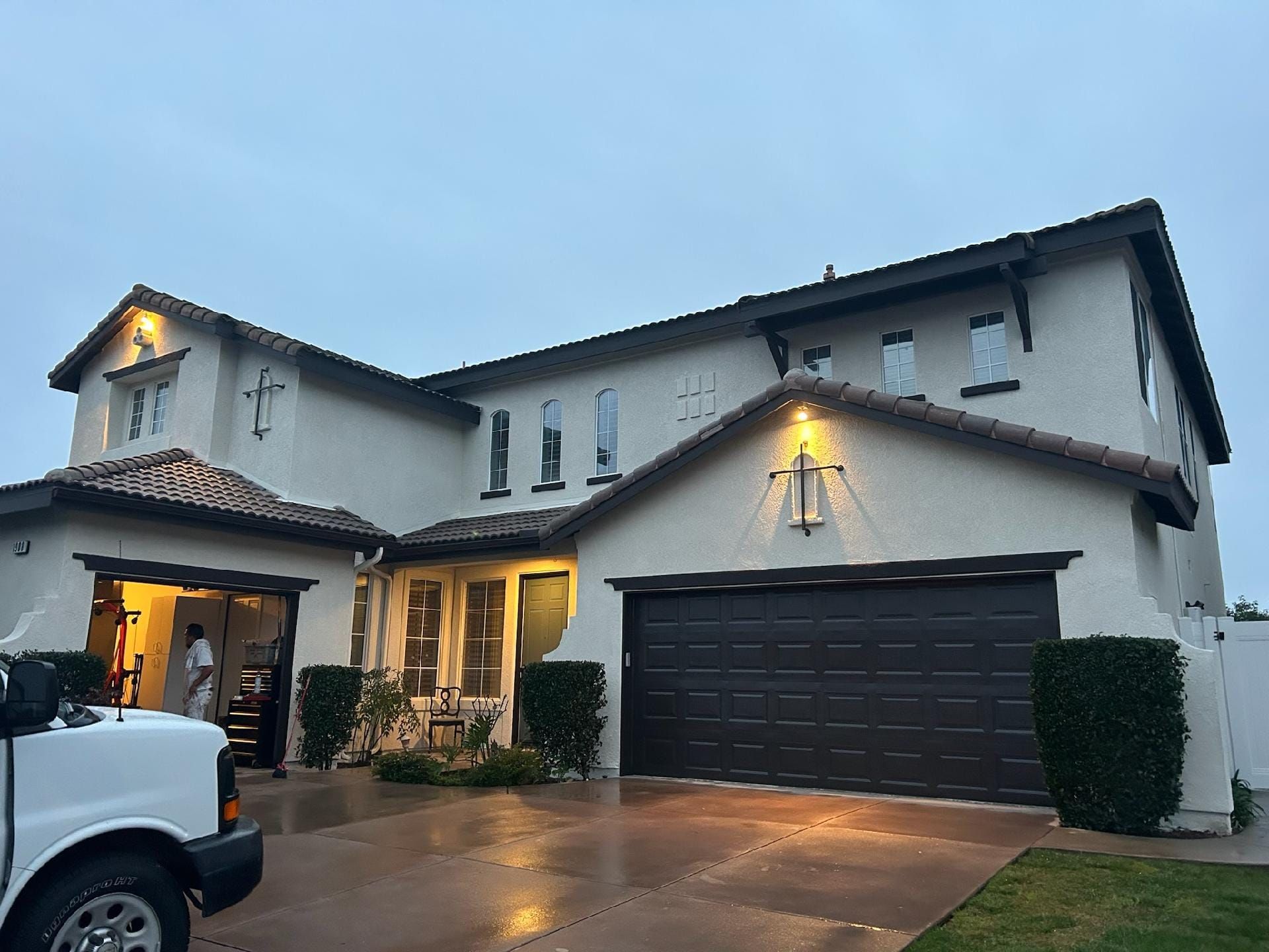 Two-story beige house with dark brown garage door, small bushes, and a white van.