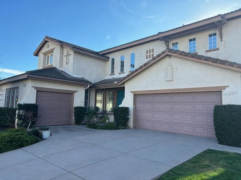 Beige two-story house with brown garage doors, a driveway, and a blue sky background.