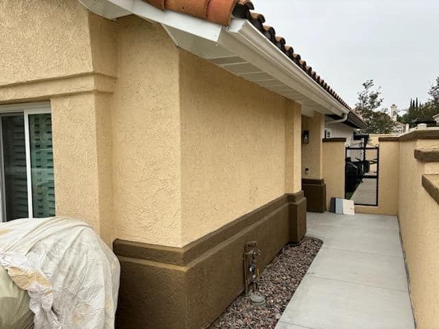 Tan stucco building with dark brown base, concrete path, gravel, and gate.