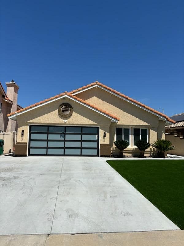 Tan house with a modern glass garage door, blue sky, green turf and concrete driveway.