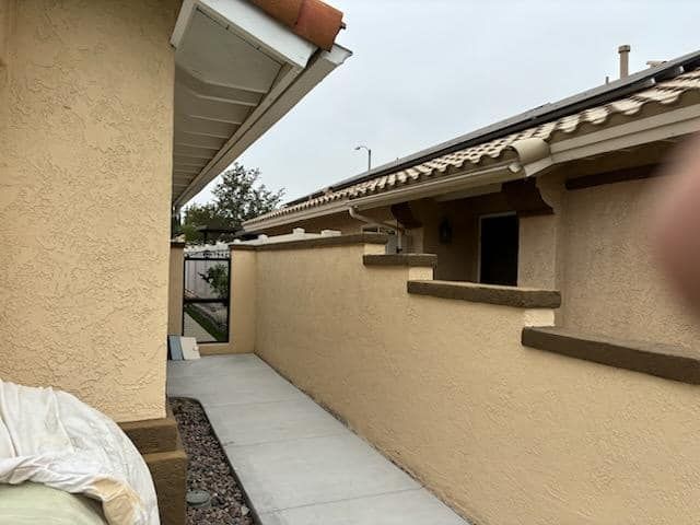 Narrow concrete path between tan walls, leading to a black gate. Overcast sky.