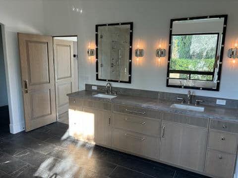 Bathroom with double vanity, two mirrors, sconce lights, and a light-colored wood cabinet with stone countertop.