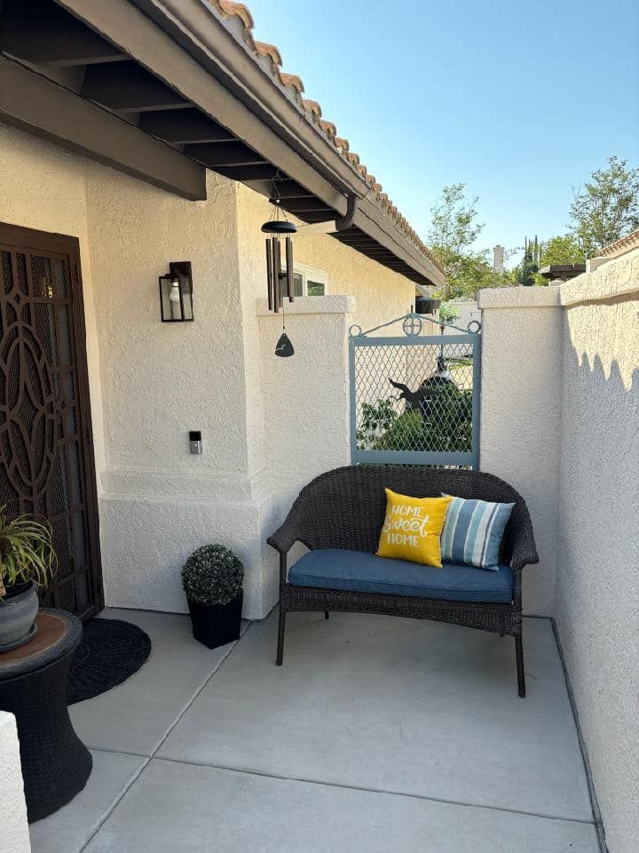 Wicker bench with blue cushion and pillows in a courtyard. Gate to a yard, potted plants, and front door.